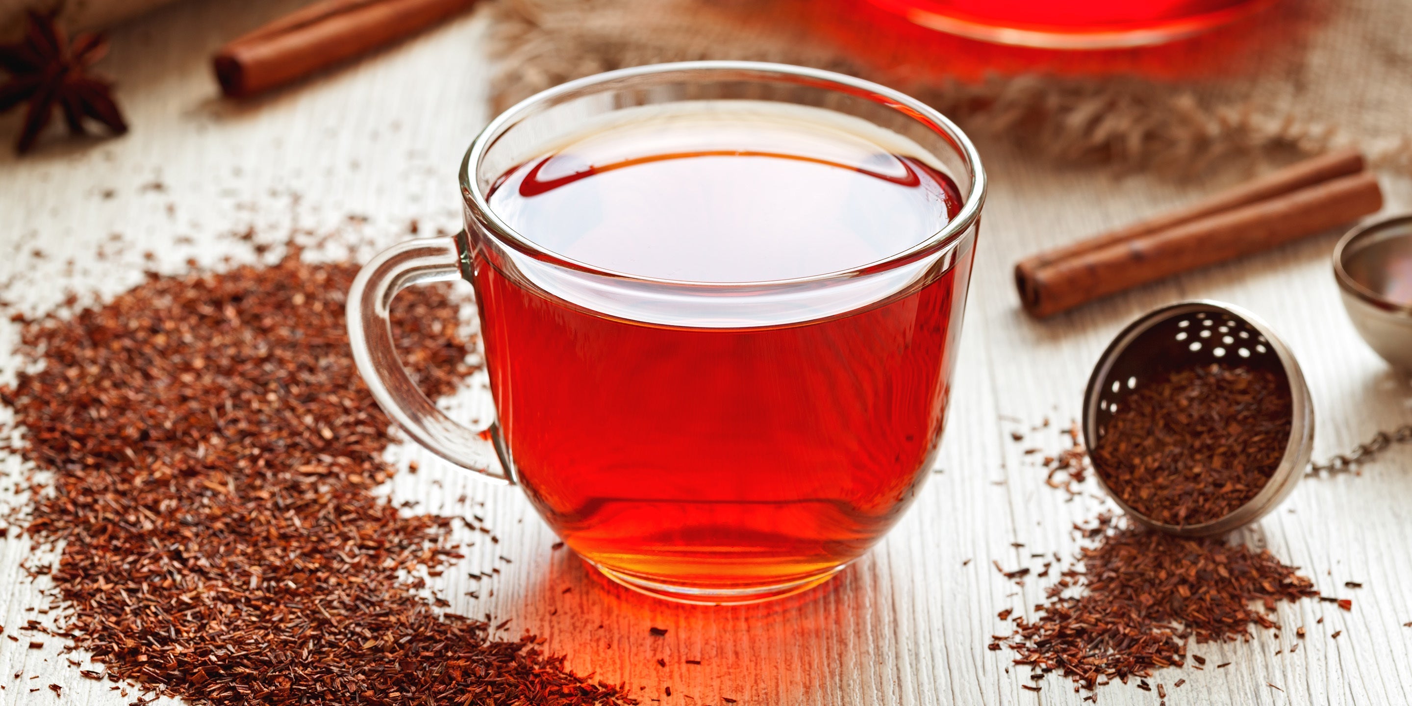 This image showcases a warm and inviting scene centered around a cup of rooibos tea. The focal point is a clear glass mug, filled with a vibrant reddish-orange liquid – the brewed rooibos tea itself. Steam gently rises from the surface, hinting at its warmth. The mug has a simple, curved handle, and is positioned slightly off-center on a light-colored, textured wooden surface. Surrounding the mug are scattered heaps of dried rooibos tea leaves, displaying their fine, reddish-brown texture. To the right of the mug, a small, perforated metal tea infuser is filled with more rooibos, suggesting the brewing process. Several cinnamon sticks lie nearby, adding a touch of spice and warmth to the composition. A piece of rough, burlap-like fabric is draped in the background, providing a rustic backdrop. A star anise is also visible, adding another aromatic element. The lighting is soft and natural, highlighting the rich colors of the tea and spices. The overall impression is one of comfort, relaxation, and a focus on natural, healthy beverages. The arrangement feels deliberately styled, emphasizing the ingredients and the ritual of tea preparation.