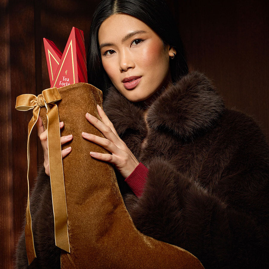 A woman in a dark brown fur coat holds a large brown suede Christmas stocking decorated with a gold ribbon and a red tea tree peeking out from the top, set against a dark wood background.