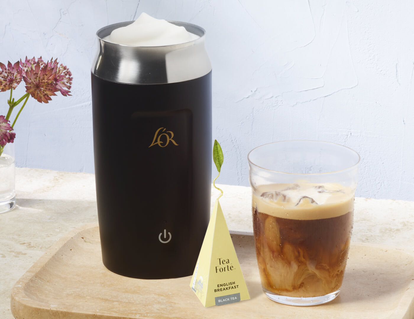 Black milk frother with a glass of iced coffee and a tea bag on a wooden board.