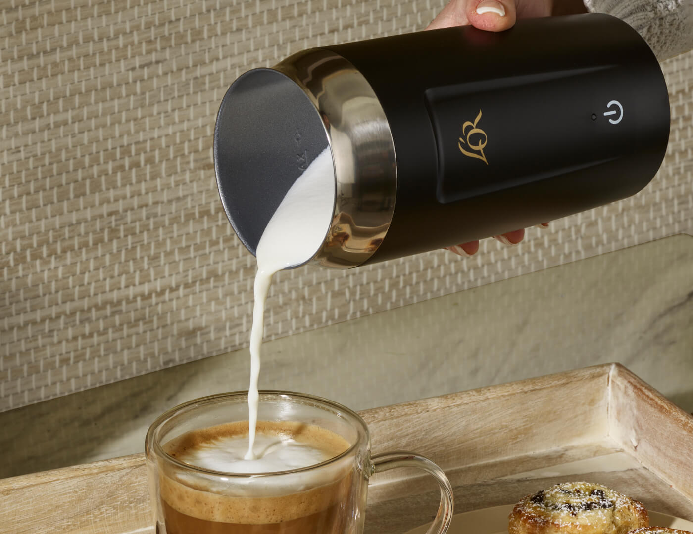Black milk frother pouring milk into a glass of coffee on a wooden tray.