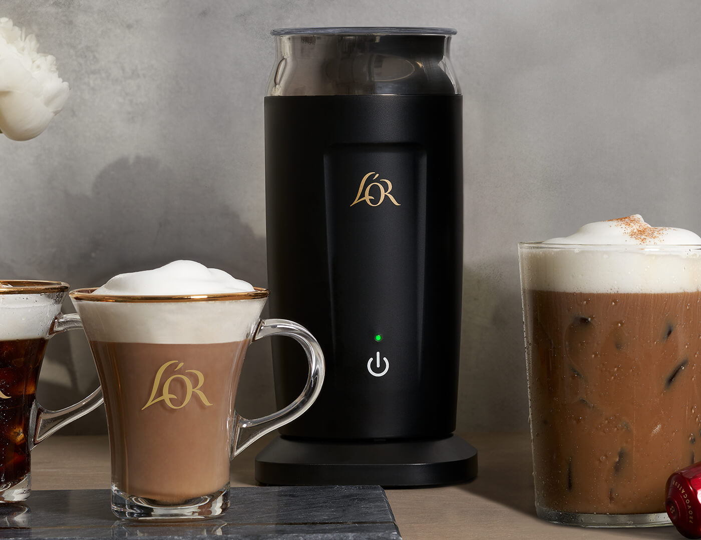 Black coffee milk frother with L&R logo next to tea and coffee drinks on a gray background