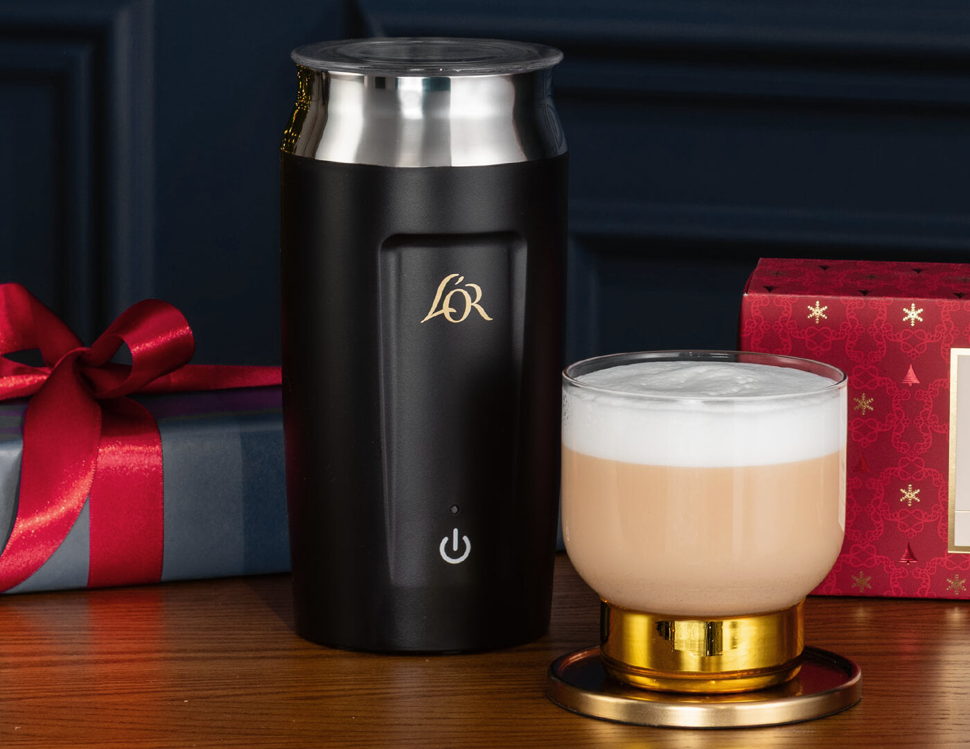 Black milk frother with a tea latte on a wooden surface with holiday tea gift boxes in the background.