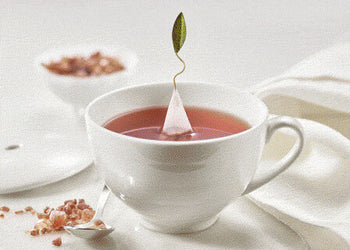White teacup with a pyramid infuser steeping in a light pink liquid on a white surface.