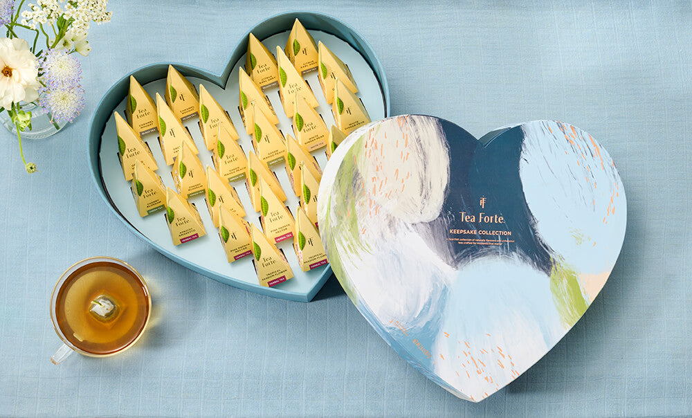 Keepsake Collection heart-shaped box of 26Tea Forté pyramid tea infusers on a light blue surface with a cup of tea.