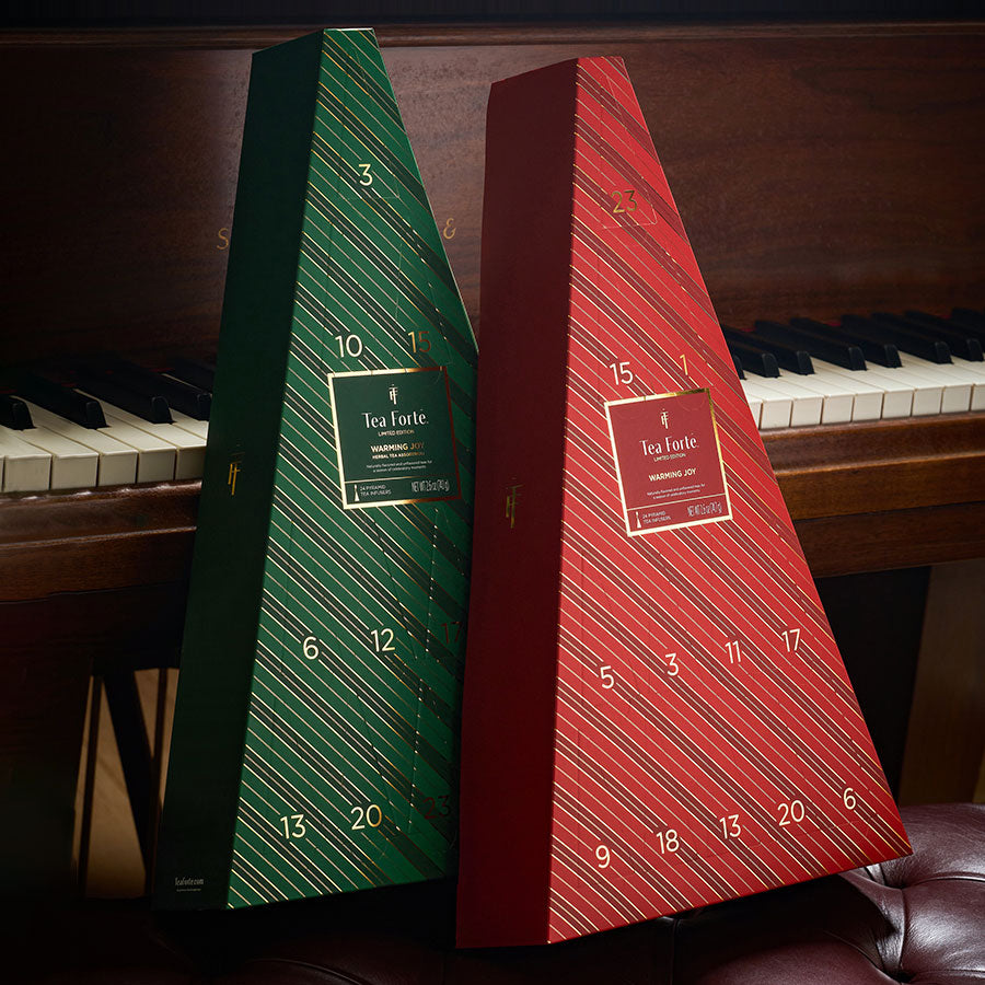 Two triangular Tea Advent Calendars with diagonal striped patterns, one green and one red, leaning against a piano. Each box has a gold label and numbered compartments on the front.