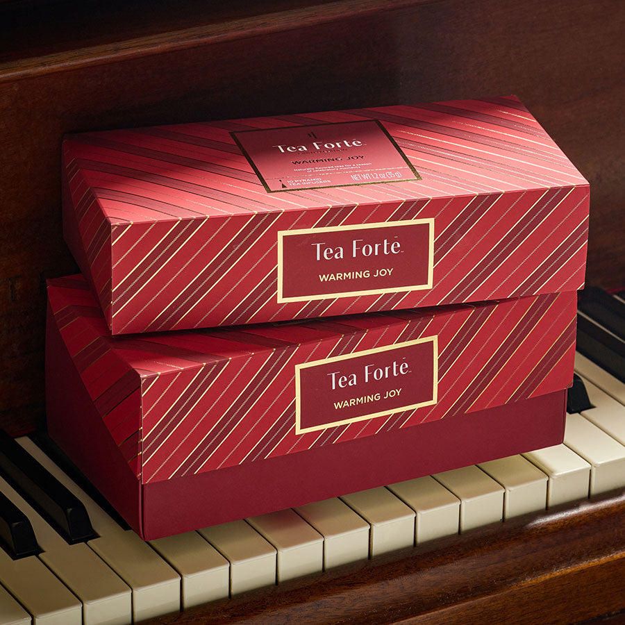 Two red Tea Forte Warming Joy Presentation Boxes of 20 teas each stacked on a piano keyboard. The boxes have a diagonal striped pattern in red and gold.