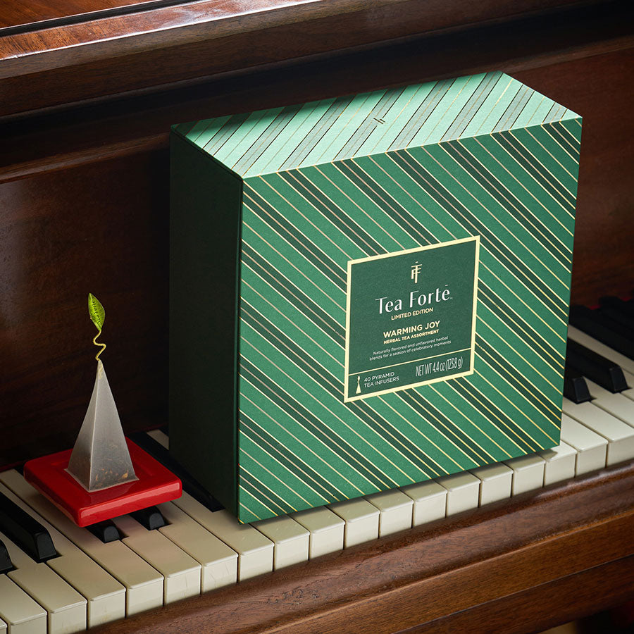 A green Tea Chest with diagonal striped pattern  placed on piano keys next to a wooden piano with a tea infuser on a red tray next to it.