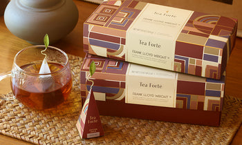 Frank Lloyd Wright - Awaken creativity with this distinct collection of grounding teas | 2 boxes stacked with glass teacup and pyramid infuser on a rattan placement on a wood table