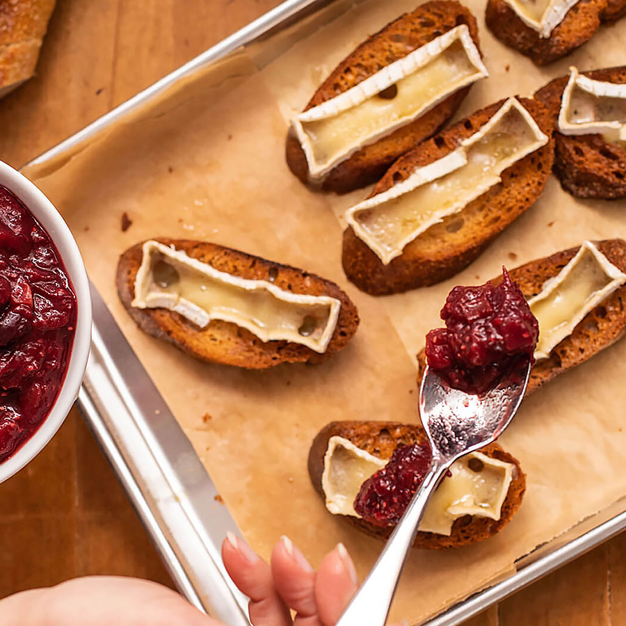 Crostini on a baking tray with cranberry topping getting spooned on