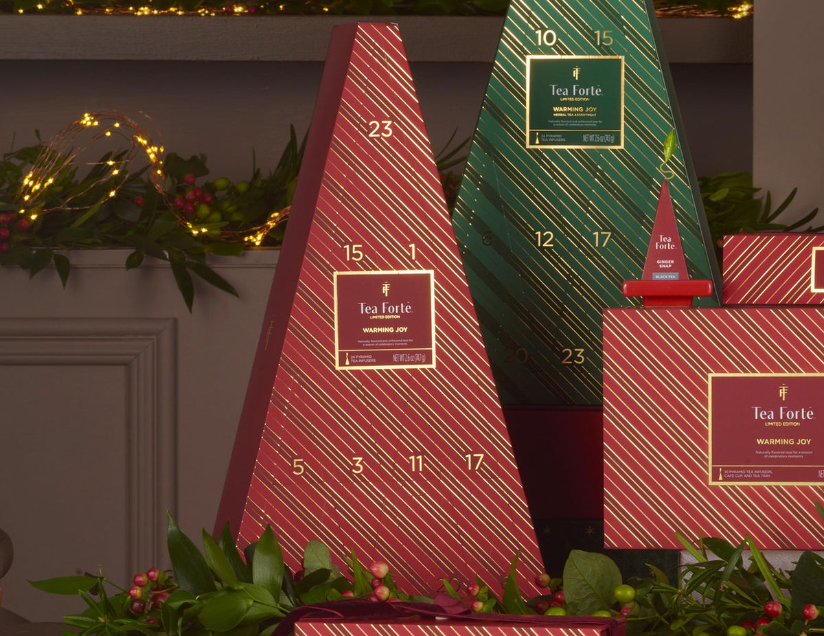 Tea Forté Christmas Advent calendar boxes arranged on a mantel with holiday greenery and warm string lights in the background. The set includes red and green tree-shaped boxes with gold diagonal stripes and numbered compartments.