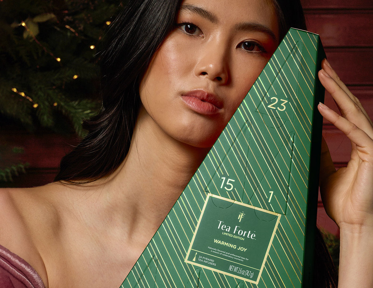 MAIN PDP | A green triangular Advent Calendar with diagonal striped pattern and gold numbers, being held by a woman in a pink dress off the shoulder, shown close to her face