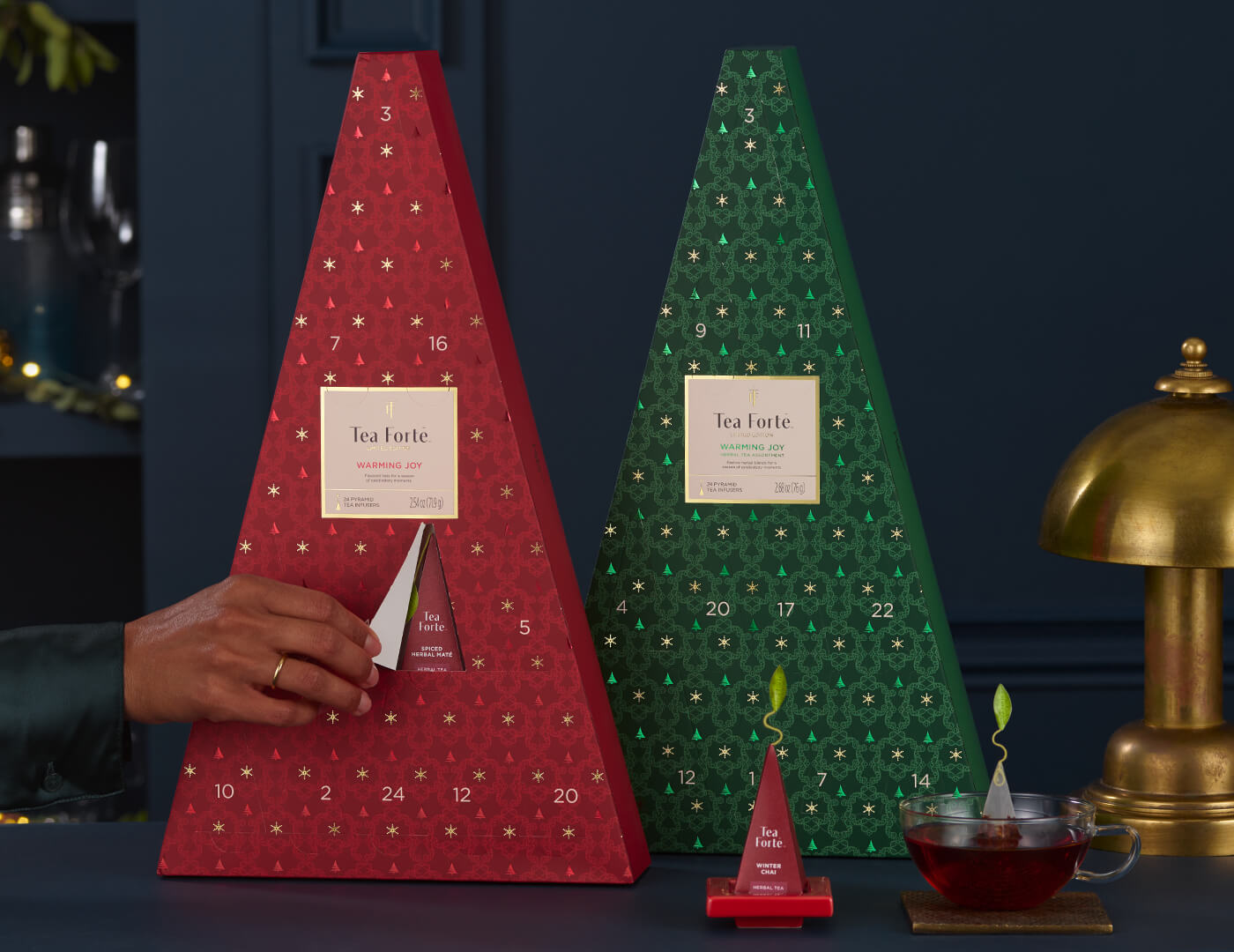 Two triangular Advent Tea Calendars, one red, one green,  with numbered doors, and a hand opening a door revealing a tea pyramid inside