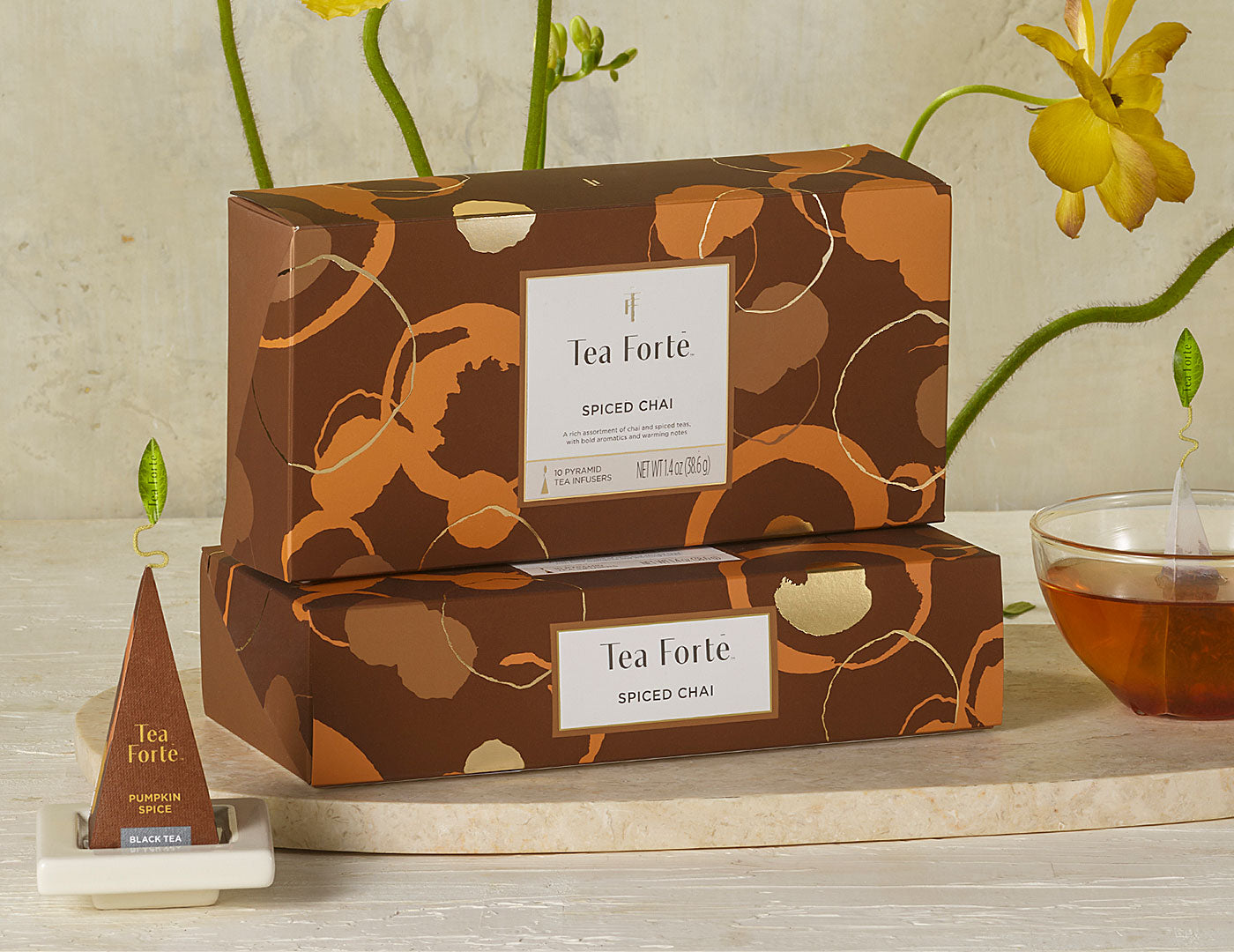 MAIN PDP | 2 Spiced Chai tea assortment boxes in 10 count petites with lid closed, infuser and cup of steeped tea to the side with yellow flowers