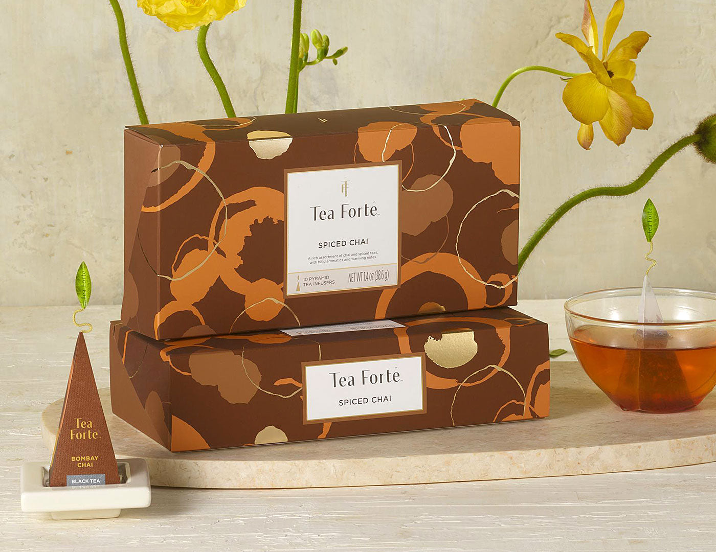 MAIN PDP | 2 Spiced Chai tea assortment boxes in 10 count petites with lid closed, infuser and cup of steeped tea to the side with yellow flowers