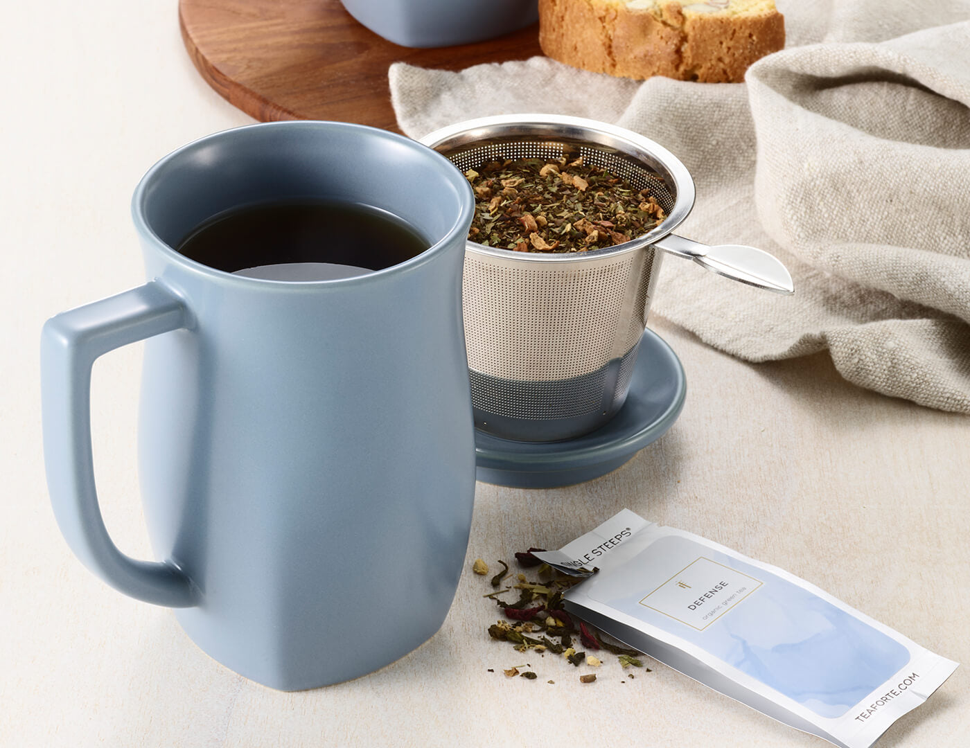 Fiore Steeping Cup and infuser with Single Steep pouch in Stone Blue on a table with tea and croissants