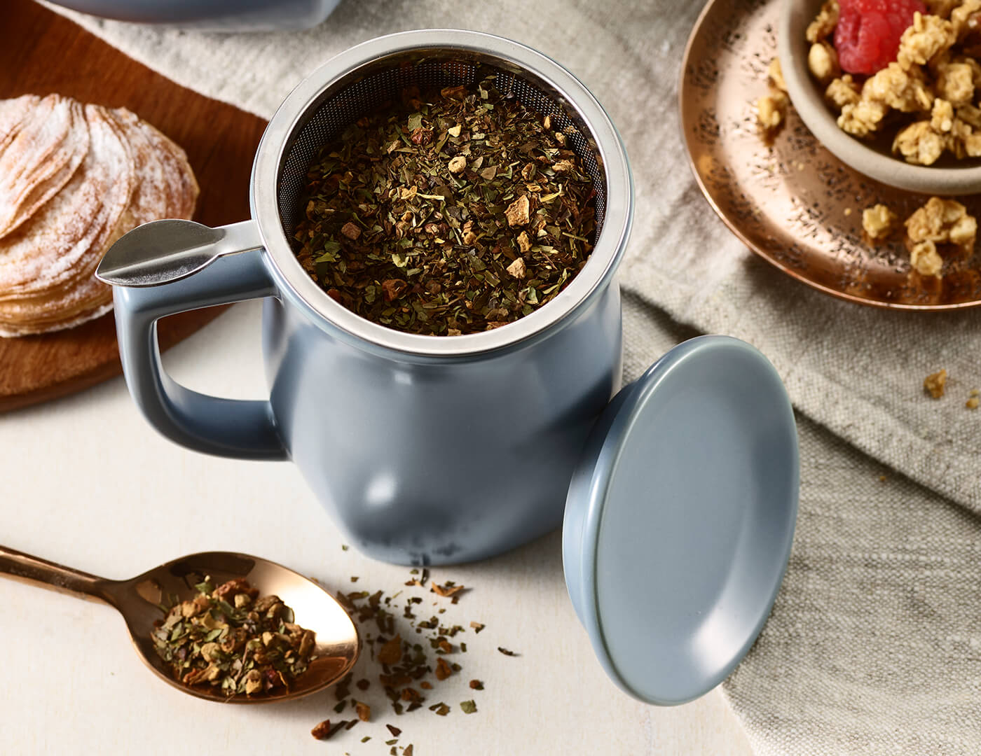 Fiore Steeping Cup and infuser in Stone Blue, open lid, on a table with tea and croissants