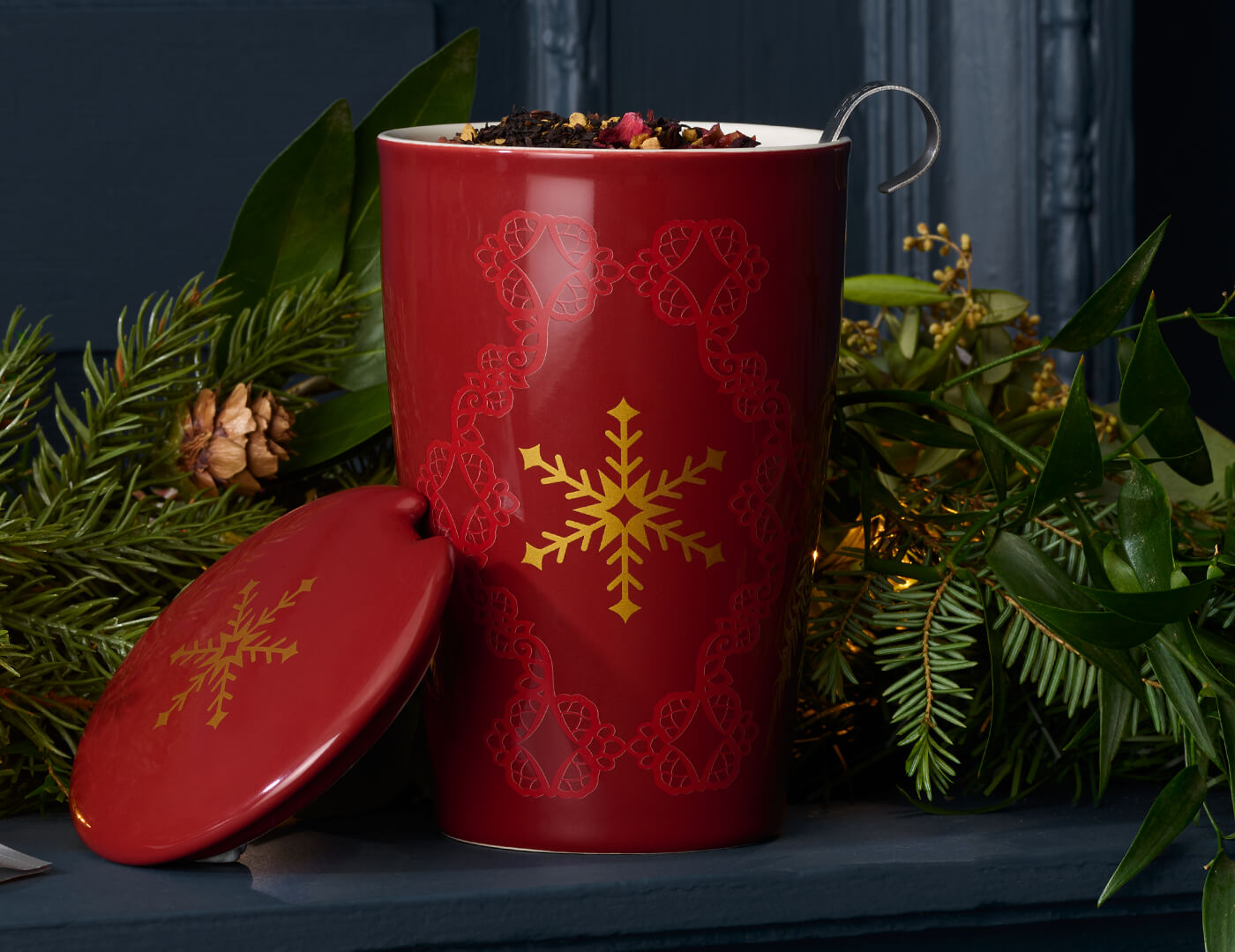 KATI® Steeping Cup & Infuser Warming Joy with loose tea inide, lid to the side, evergreen background on a shelf