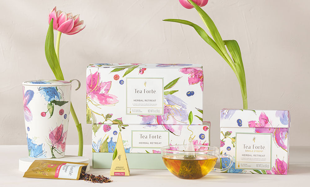 Herbal Retreat tea assortment in various boxes on table with a pyramid tea infuser,  teacups, pouch of loose leaf tea and pink tulips.