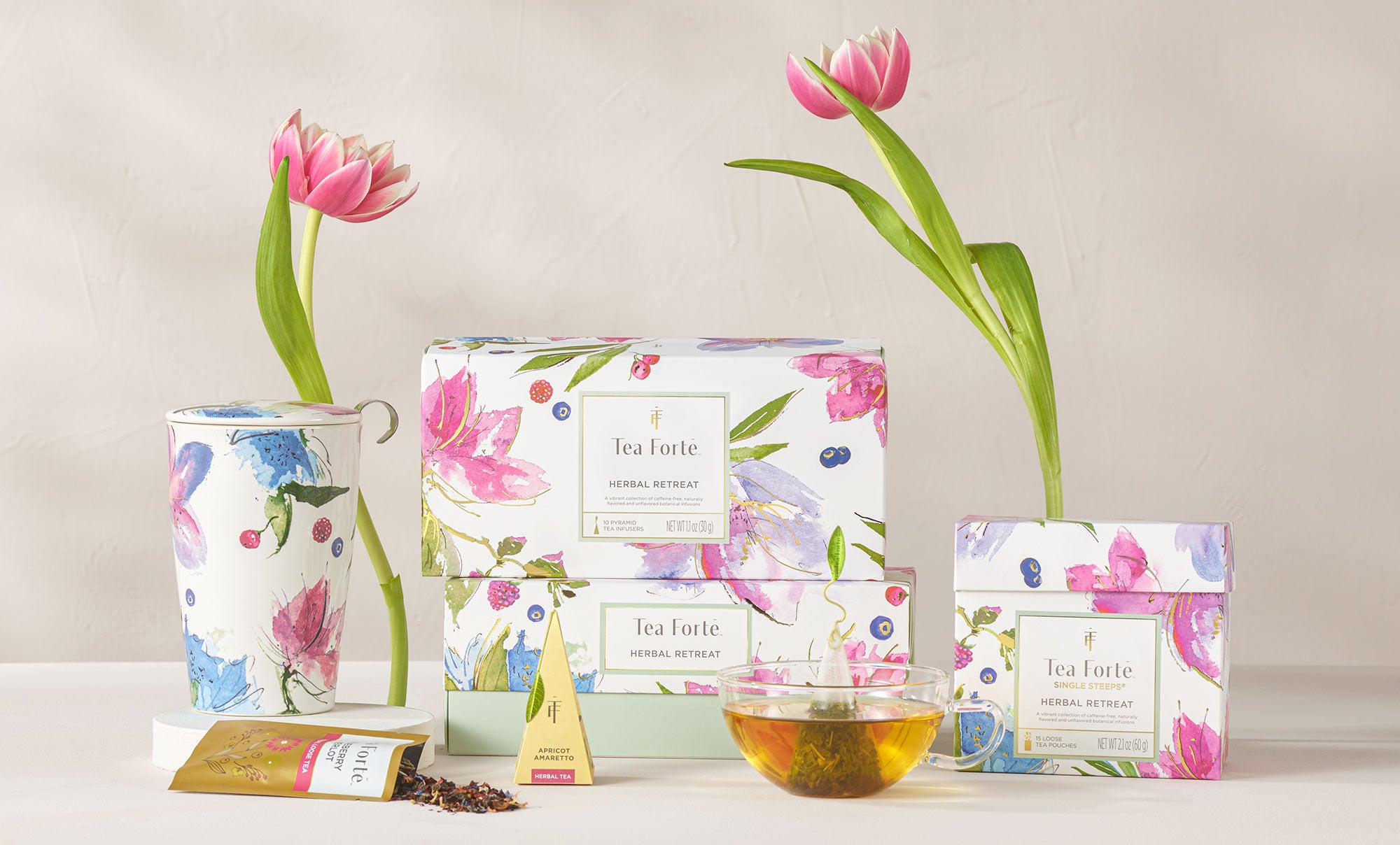 Various boxes of Herbal Retreat colection with teacups, a pyramid infuser, a pouch of loose leaf tea and pink tulips.