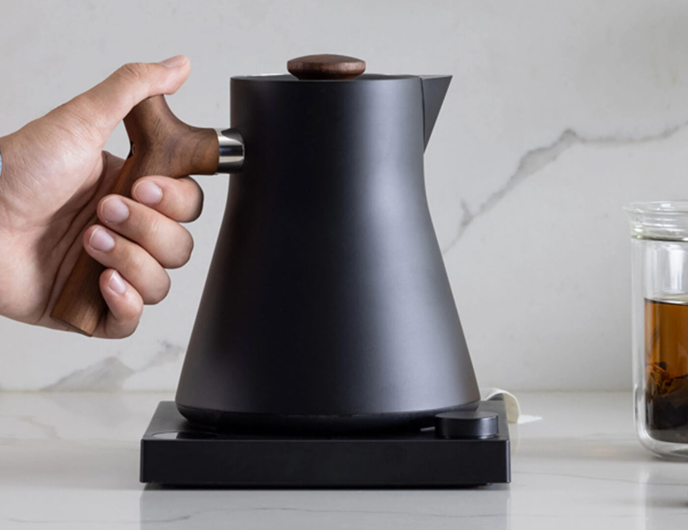 Hand on the handle of a Black Corvo Kettle EKG with Walnut handle, resting on the base