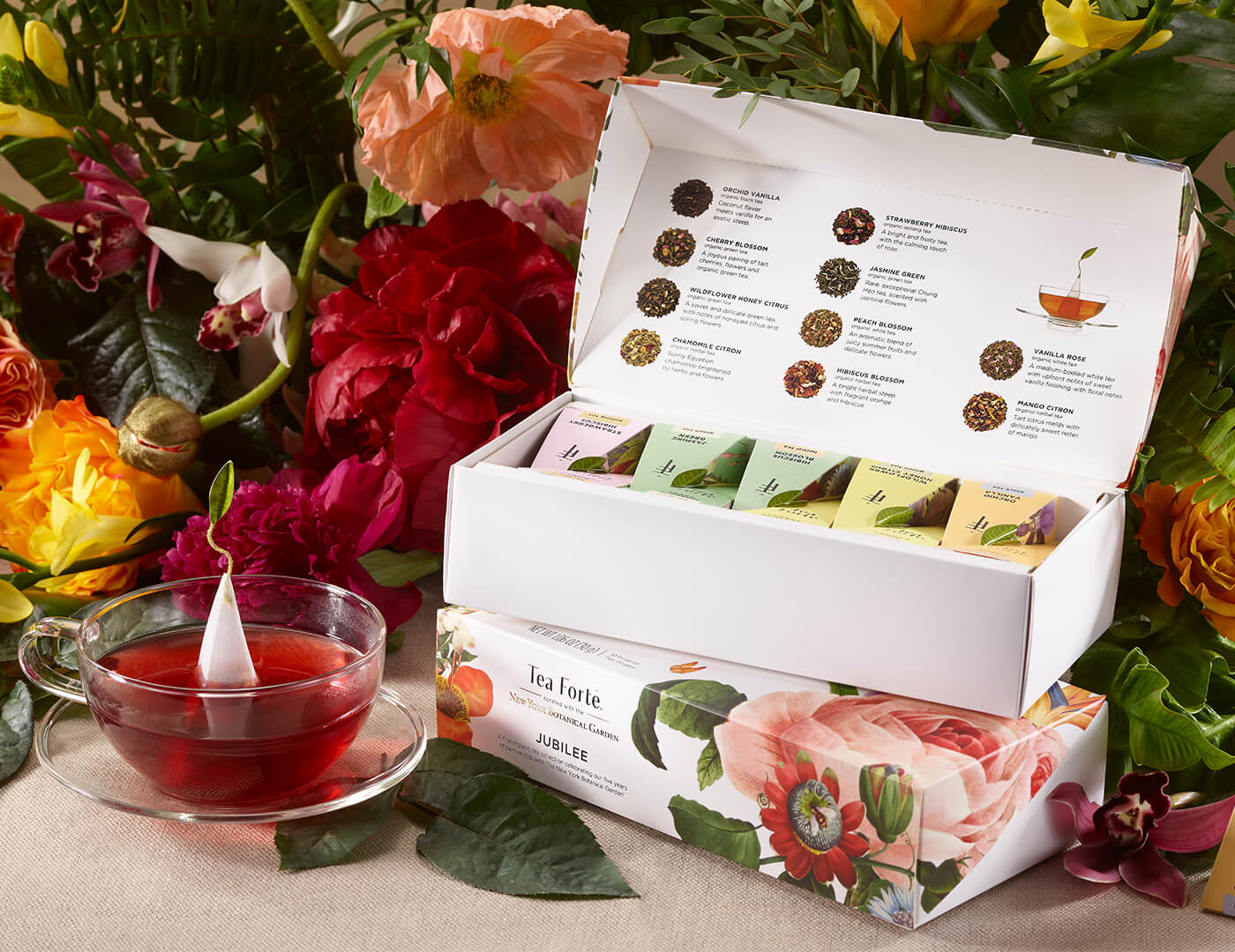 Jubilee Petite Presentation Box of 10 pyramid teas, lid open, on a table with flowers and a glass cup of tea
