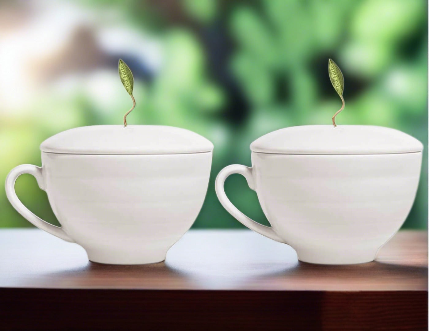 Café Cup Duo, 2 white teacups with lids on and pyramid infuser leaves poking out of the hole in the top of each lid on a table with a green foliage in the background
